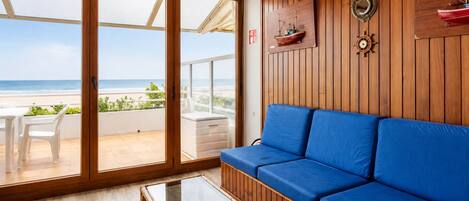 A room with a wooden wall, a blue sofa, a glass table, and a view of the beach.