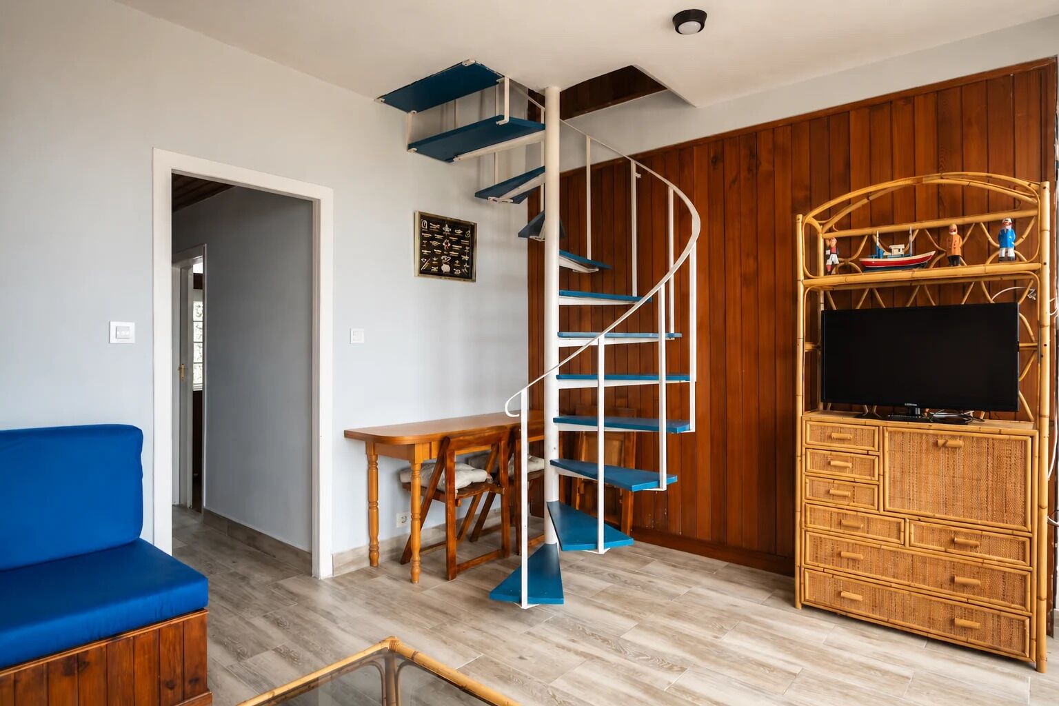 Una sala de estar con un sofá azul, una mesa de centro de madera, una escalera en espiral, un televisor sobre un mueble de bambú y una pared de madera.