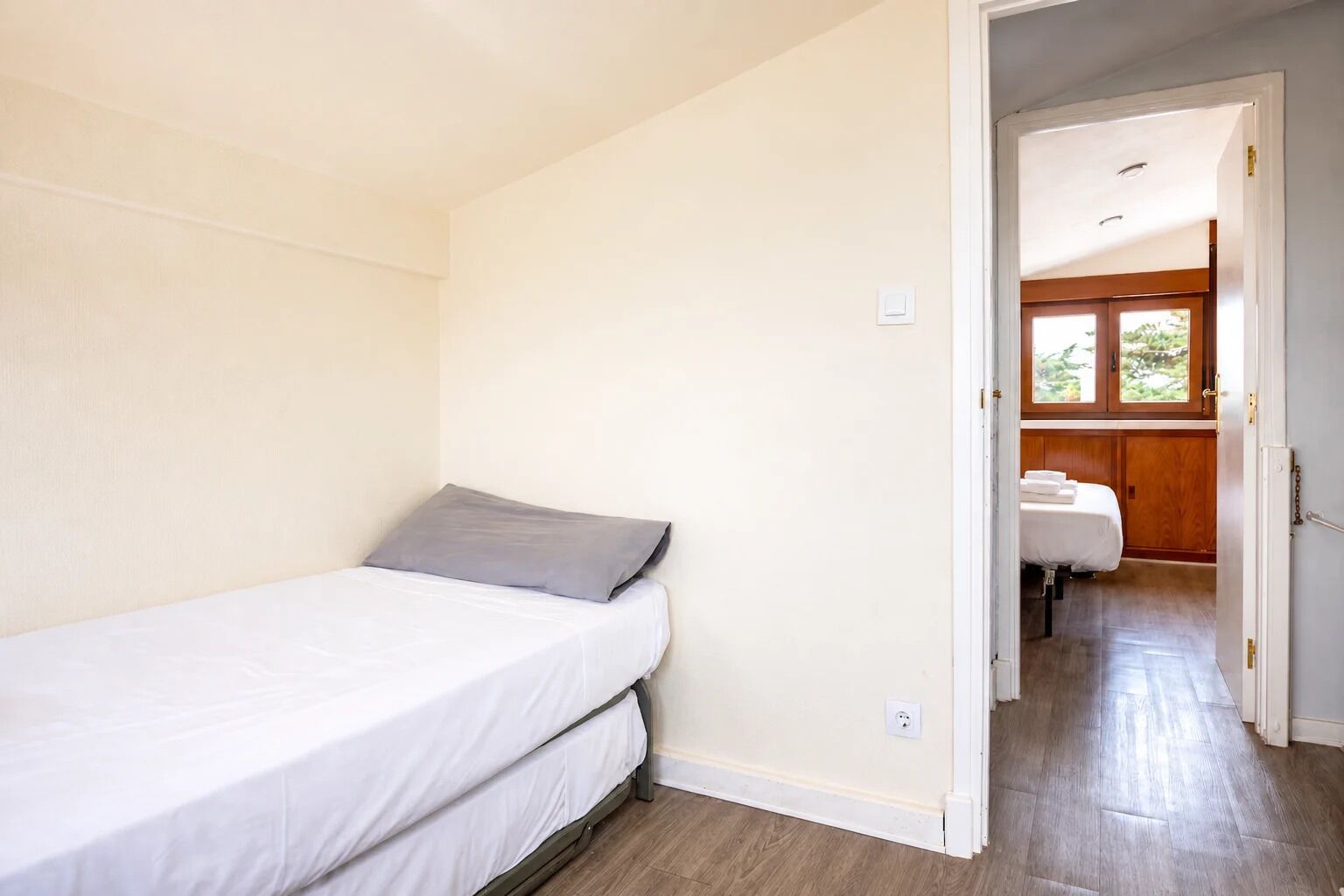 Quarto individual com uma cama, piso de madeira, parede branca e porta que dá para um banheiro com armário de madeira.
