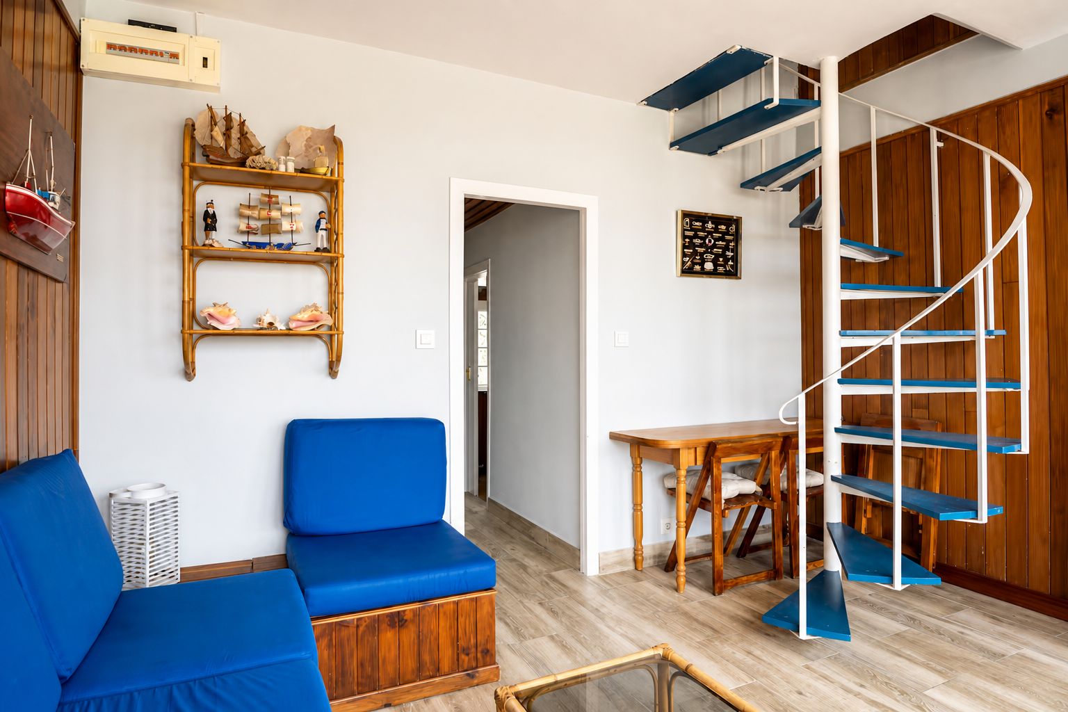 Una habitación con sofá azul, una mesa de madera, una escalera de caracol y un estante empotrado con objetos decorativos.