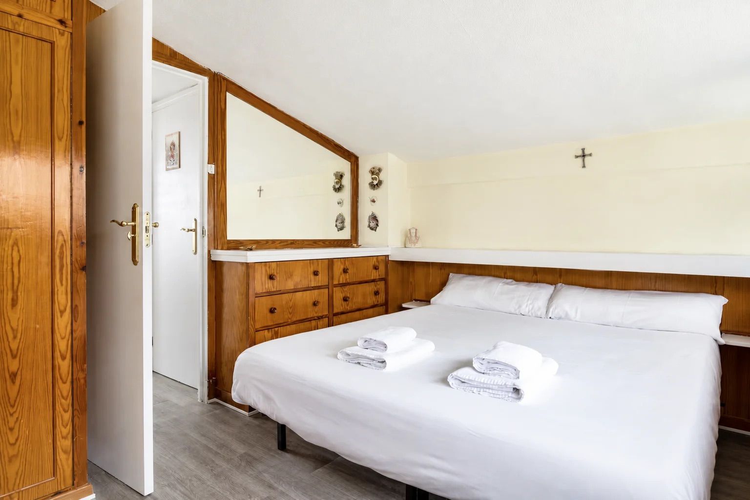 Habitación de hotel con cama, armario de madera, espejo y una cruz en la pared.