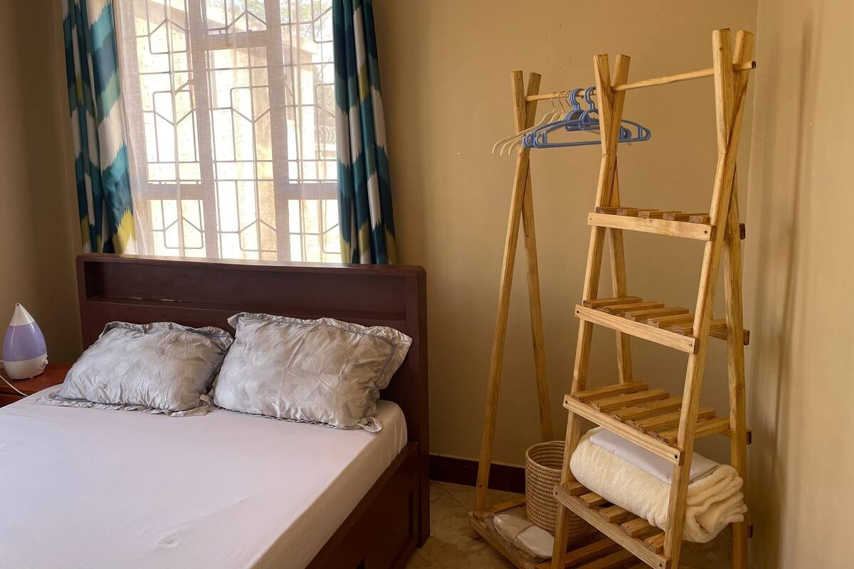 A bed with two pillows, a wooden shelf unit with hangers, and a window with patterned curtains.