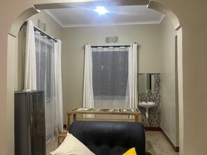 Habitació amb un sofà negre, una taula de fusta, un frigorífic, un rentaplat i una finestra amb cortines.
