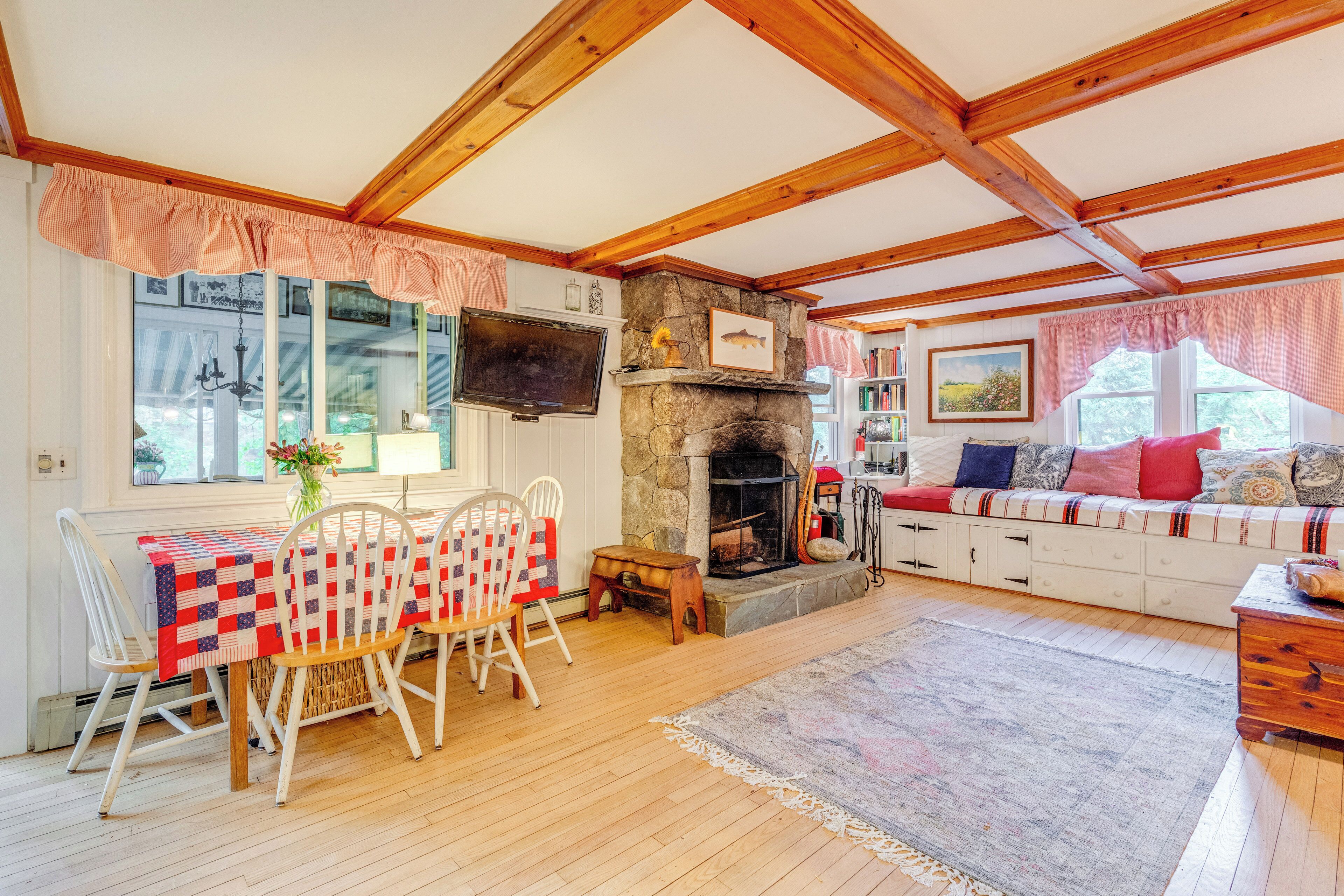 Una sala de estar acogedora con chimenea de piedra, vigas de madera, un comedor con una mesa cubierta con colcha y una zona de estar confortable con cojines.
