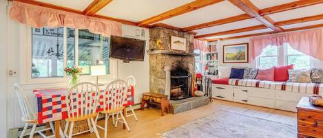 Una sala de estar acogedora con chimenea de piedra, vigas de madera, un comedor con una mesa cubierta con colcha y una zona de estar confortable con cojines.
