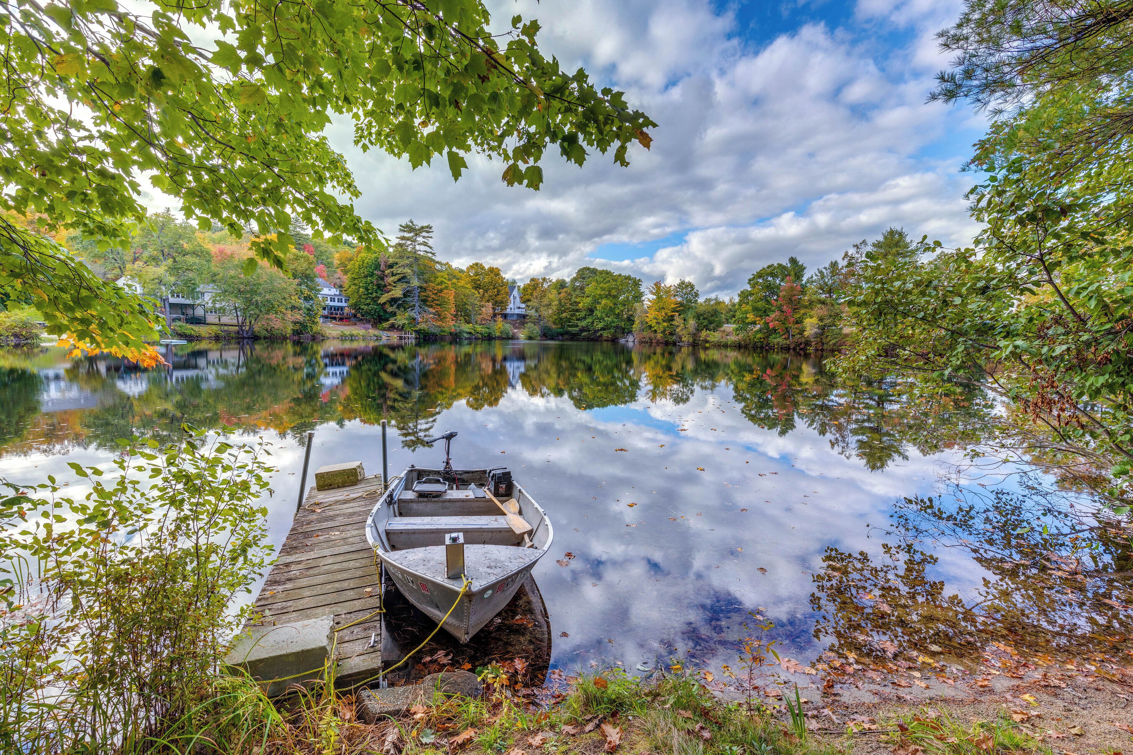 Un lago tranquilo con un muelle y un bote, rodeado de árboles y una casa al fondo.