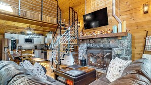 A spacious living area with a stone fireplace, a comfortable sofa set, a dining table with chairs, a kitchen in the background, and a staircase leading to an upper level.