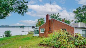 A brick house with a white garage, a grassy yard, and a lake in the background. - Webster Lake Home w/ Patio ~ 3 Mi to Town! (North Webster)