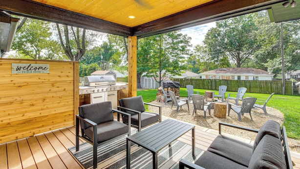 A covered patio area with outdoor furniture, a grill, and a view of a backyard with a lawn and trees. - Stylish & Modern Lenexa Haven w/ Fire Pit & Deck! (Lenexa)
