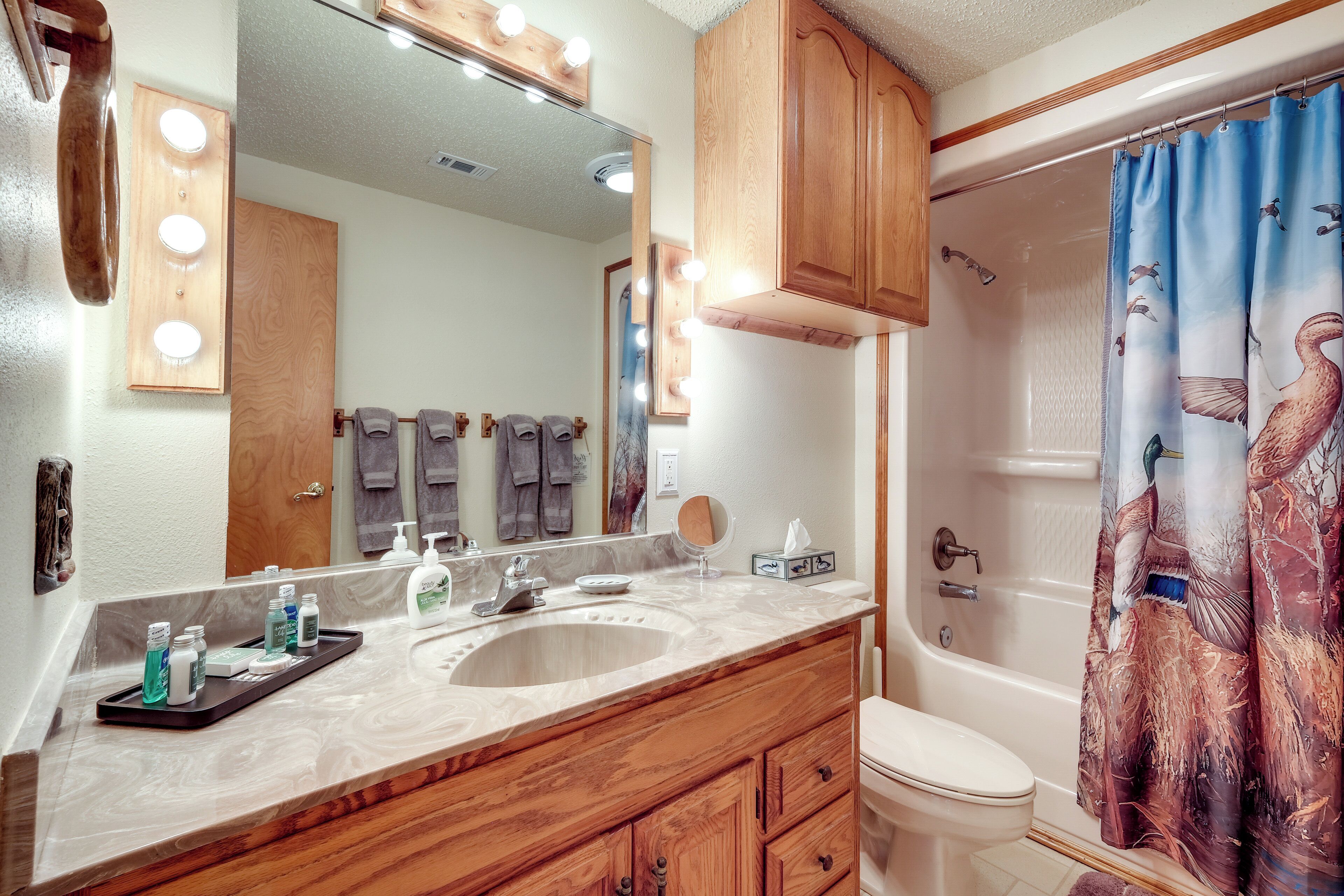 A bathroom with a wooden vanity, a large mirror with lights, a bathtub with a shower curtain, and a toilet.