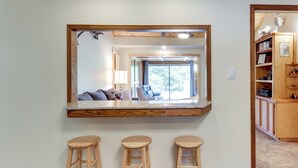 Une pièce dotée d’un grand miroir, de trois tabourets en bois, d’une bibliothèque en bois et offrant une vue sur un espace de vie comprenant un canapé et un fauteuil.