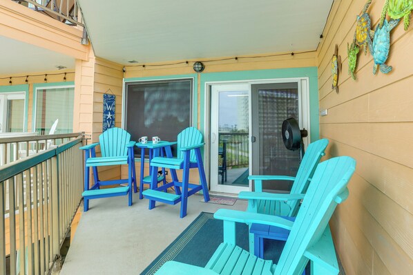 A balcony with blue Adirondack chairs, a small table, and a door leading to an outdoor area. - Walk to Seawall & Beach! Galveston Condo w/ Pool (Galveston)