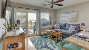 A living room with a sofa, coffee table, and a TV mounted on the wall. There is a large window with curtains, a ceiling fan, and a painting on the wall. - Walk to Seawall & Beach! Galveston Condo w/ Pool (Galveston)