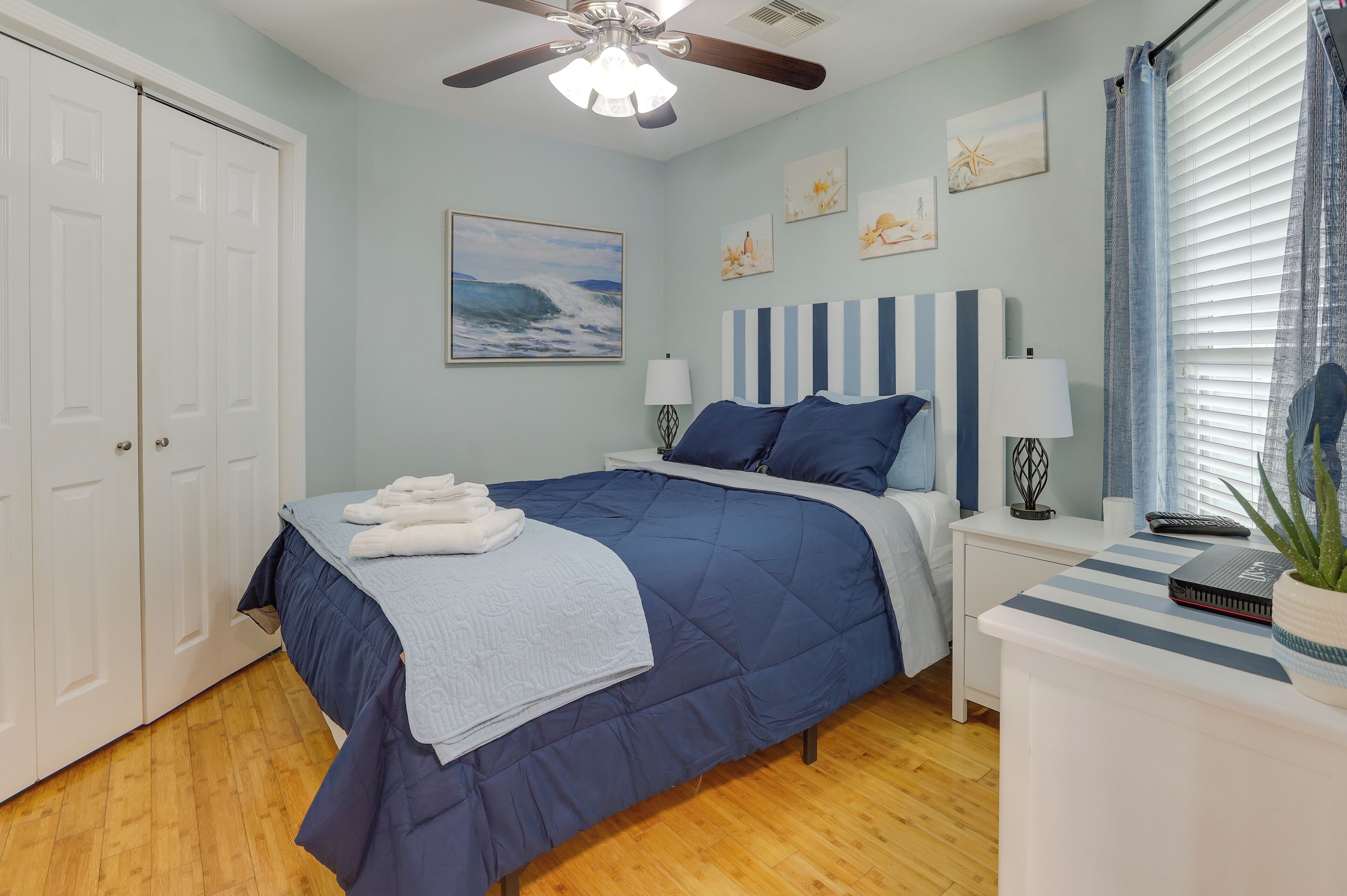A bedroom with a bed, a nightstand, a ceiling fan, a window with curtains, and artwork on the wall.