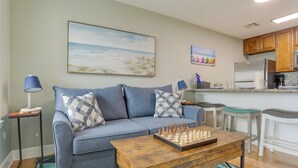 A living room with a sofa, coffee table, and a chessboard. There is a kitchen area with a refrigerator and bar stools. - Walk to Seawall & Beach! Galveston Condo w/ Pool (Galveston)
