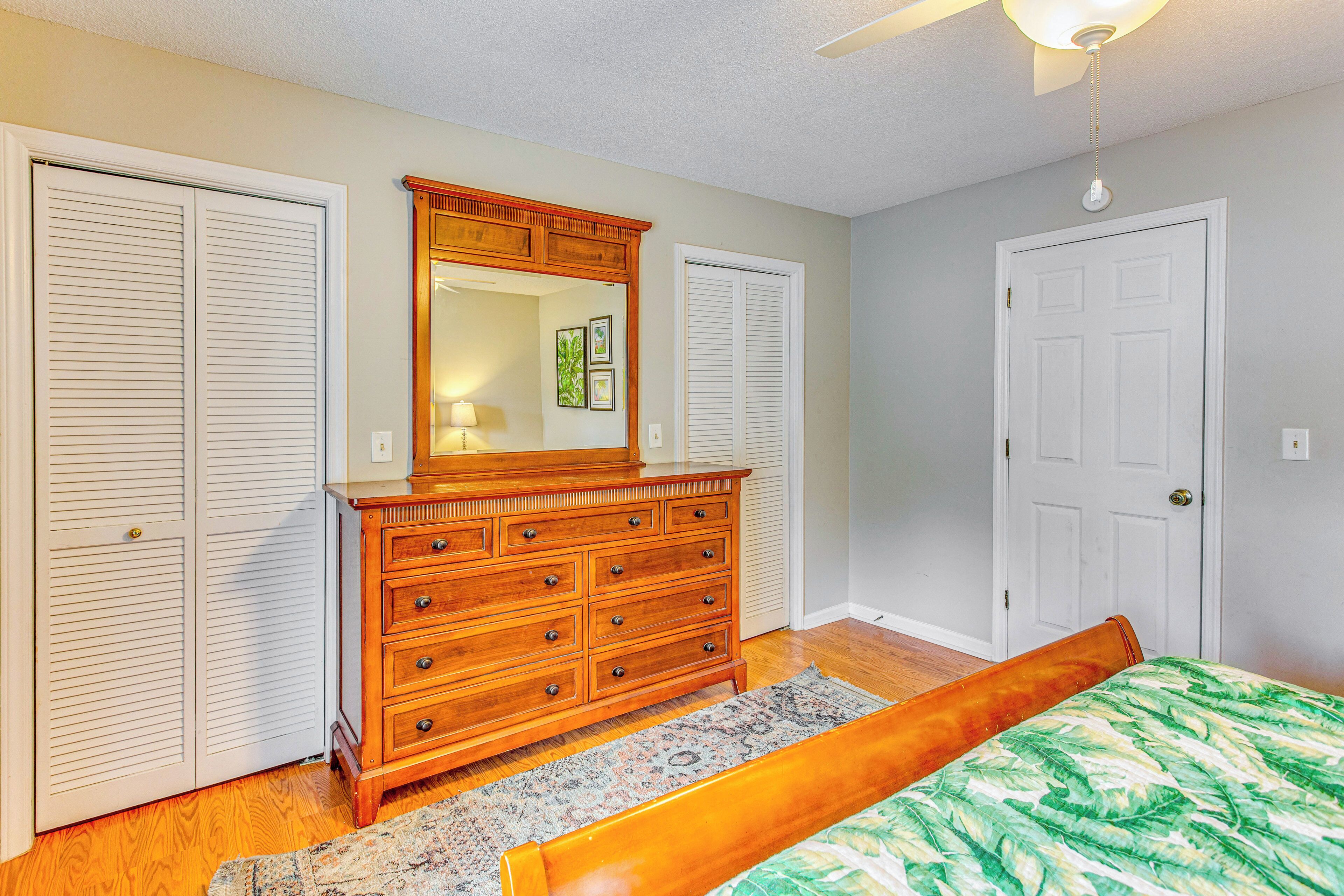 Une chambre avec une commode en bois, un lit recouvert d’une couvre-lit à motifs de feuilles, un tapis à motifs et un ventilateur de plafond.