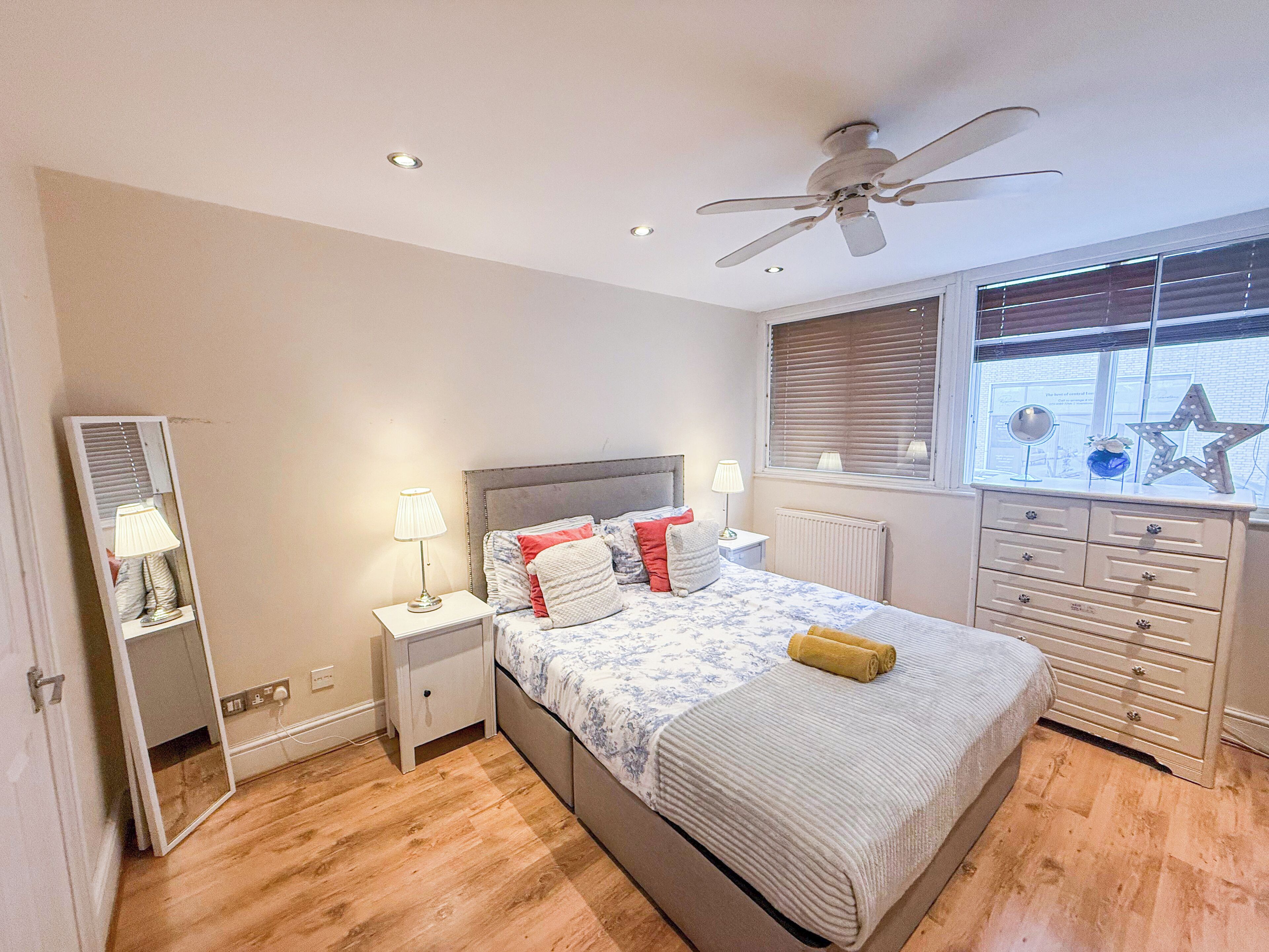 A bedroom with a bed, bedside tables, a dresser, a ceiling fan, a window with blinds, and a radiator.