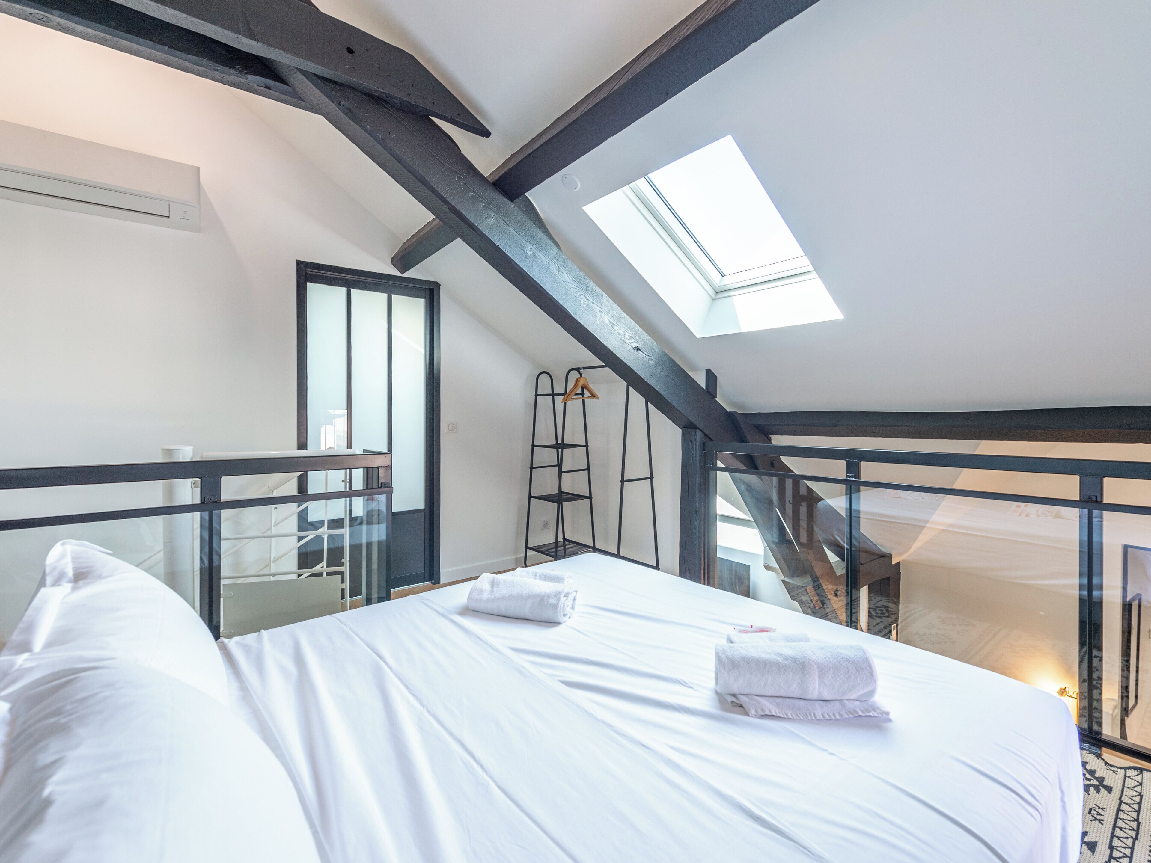 Un dormitorio con tragaluz, una cama con ropa de cama blanca, una escalera y una barandilla de vidrio.