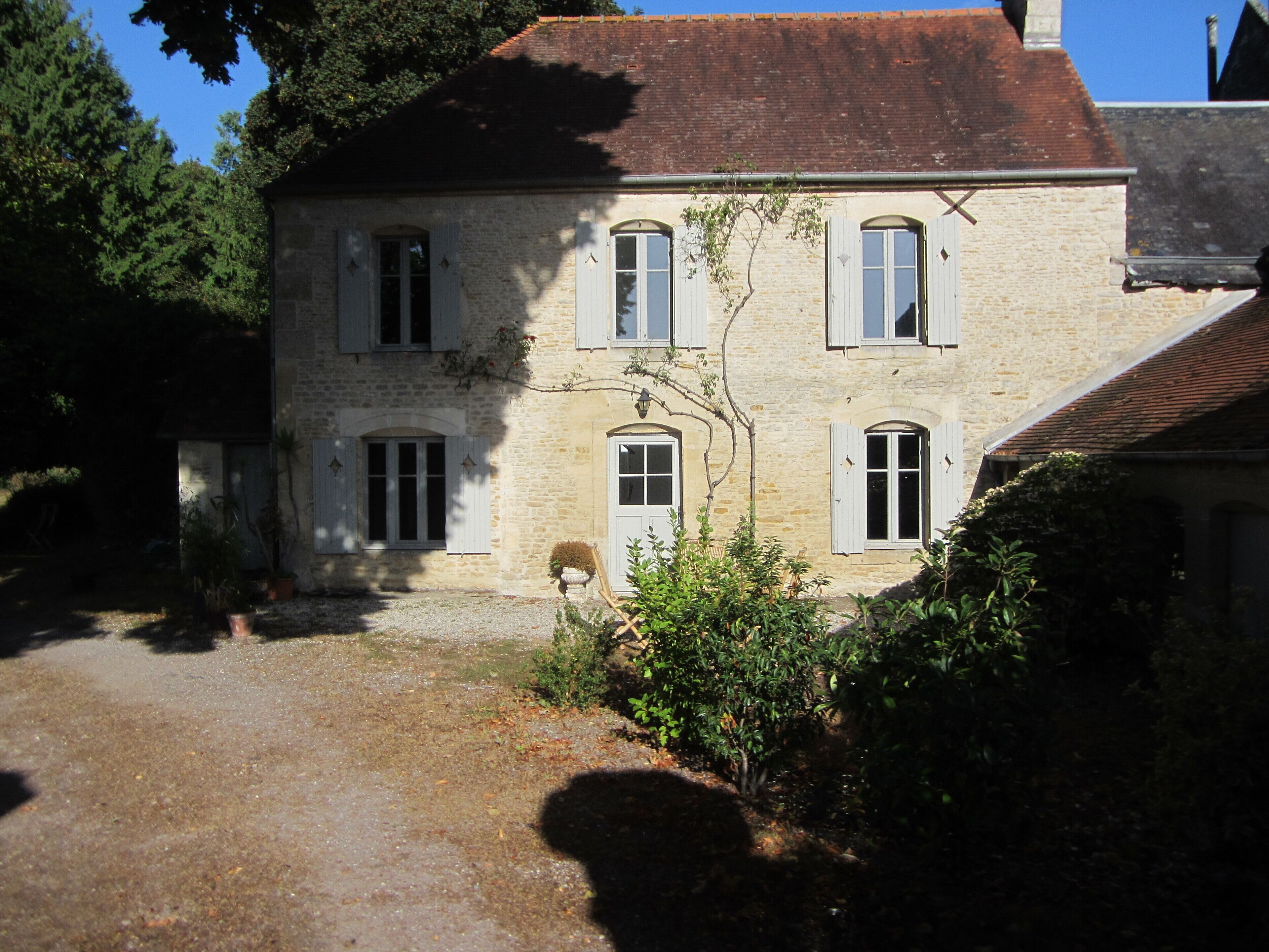 Un bâtiment en pierre doté d’une façade symétrique, de nombreuses fenêtres avec volets et d’une allée gravillonnée.