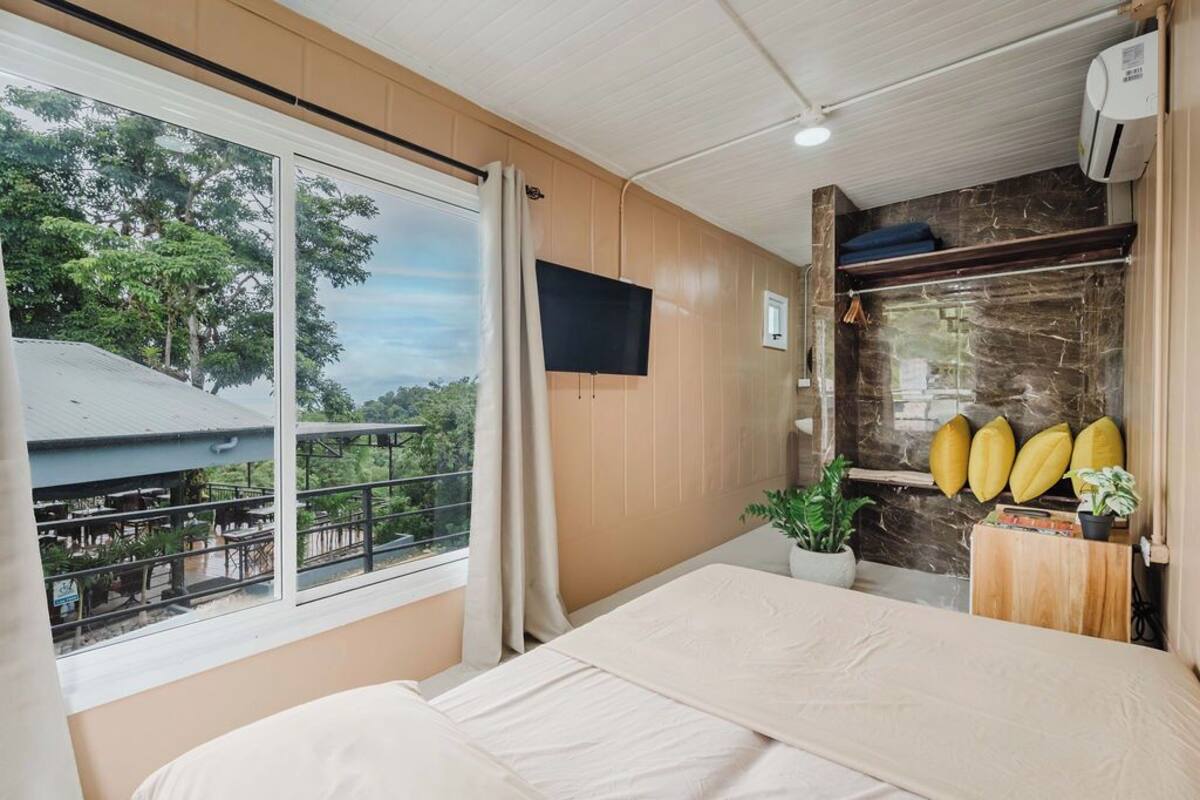 Ein Hotelzimmer mit einem großen Bett, einem an der Wand befestigten Fernseher, einem Fenster mit Blick ins Freie und einer kleinen Pflanze auf einem Holznachttisch.