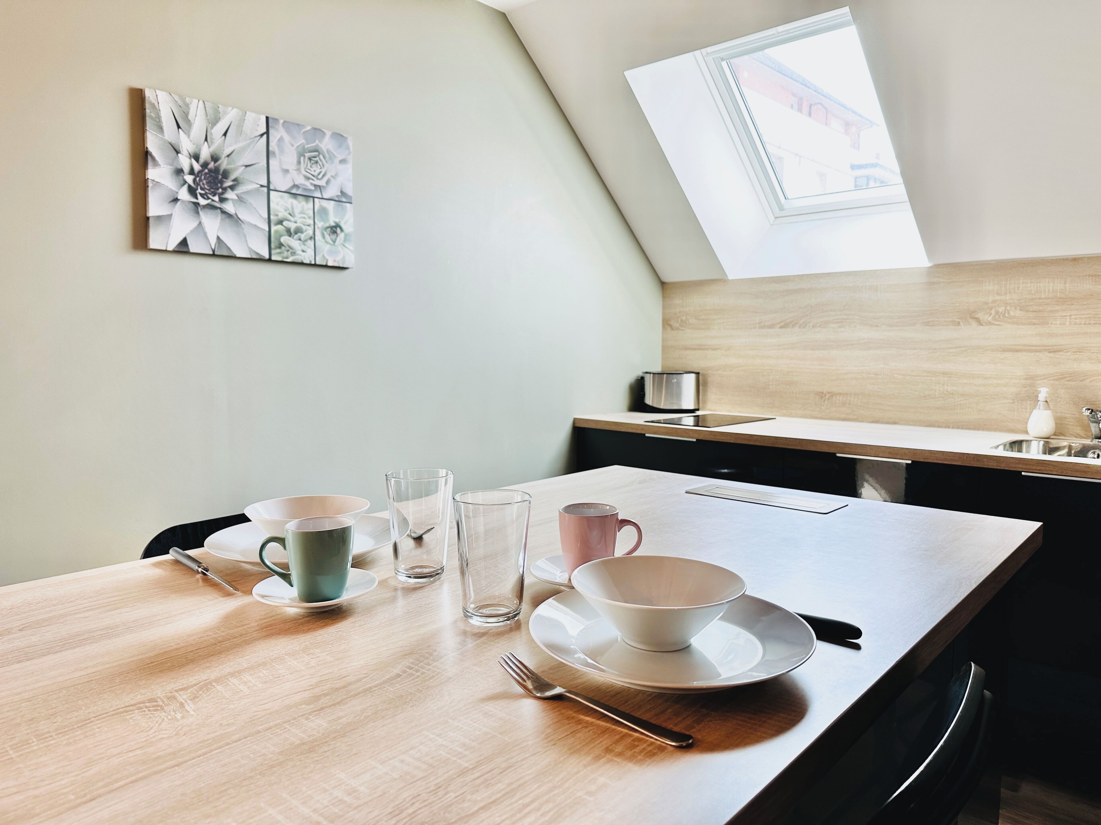 Une cuisine moderne avec un plan de travail en bois, une table à manger dressée avec de la vaisselle et des verres transparents, une fenêtre offrant une belle vue et une œuvre d’art florale accrochée au mur.