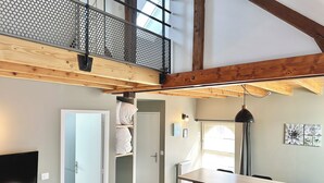 A loft-style room with a high ceiling, exposed wooden beams, a mezzanine with a metal railing, a dining area with a table and chairs, and a television mounted on the wall. - Le Patio de Loiseau  (Le Mans)