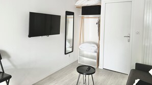 A modern room with a flat-screen TV, a full-length mirror, a wardrobe with hanging space, a small round table, and a door. - Le Patio de Loiseau  (Le Mans)