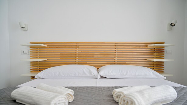 Ein ordentlich bezogenes Bett mit einem Holz-Kopfteil, weißen Kissen und zwei aufgerollten Handtüchern auf einer grauen gesteppten Bettdecke.