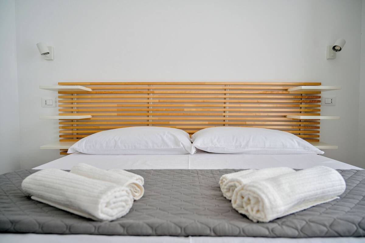 Un lit bien fait, avec une tête de lit en bois, des oreillers blancs et deux serviettes enroulées posées sur une couverture matelassée grise.