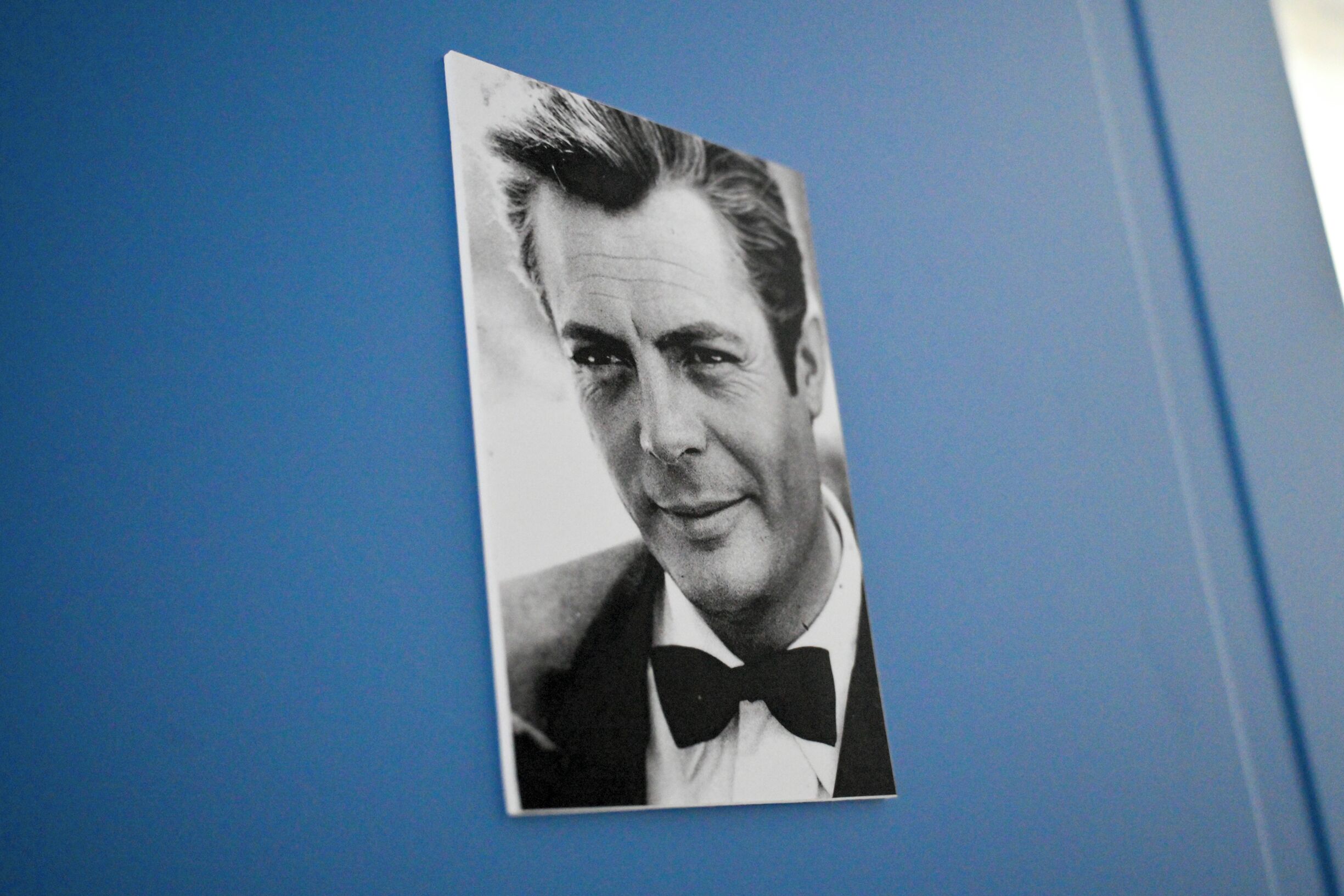 Fotografía en blanco y negro de un hombre con esmoquin y corbata de lazo, sobre un fondo azul.