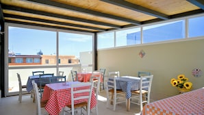 Eine sonnendurchflutete Terrasse mit Tischen und Stühlen, großen Fenstern und Blick auf Gebäude.