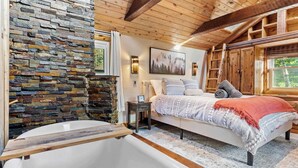 Un dormitorio con pared de piedra, una cama grande, techo de madera y una bañera.