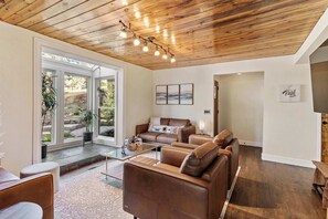 Un salon moderne avec un plafond en bois, des canapés en cuir, une table basse en verre et une vue sur un espace extérieur, accessible par de grandes portes vitrées.