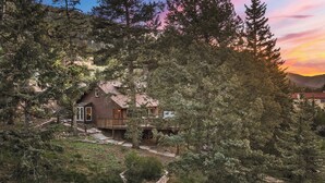 Una cabaña de troncos con terraza, rodeada de un bosque denso y montañas.