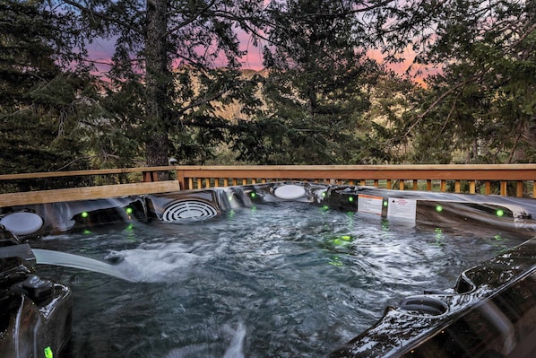 A hot tub with multiple jets surrounded by a wooden deck, set against a backdrop of trees and a sunset sky.
