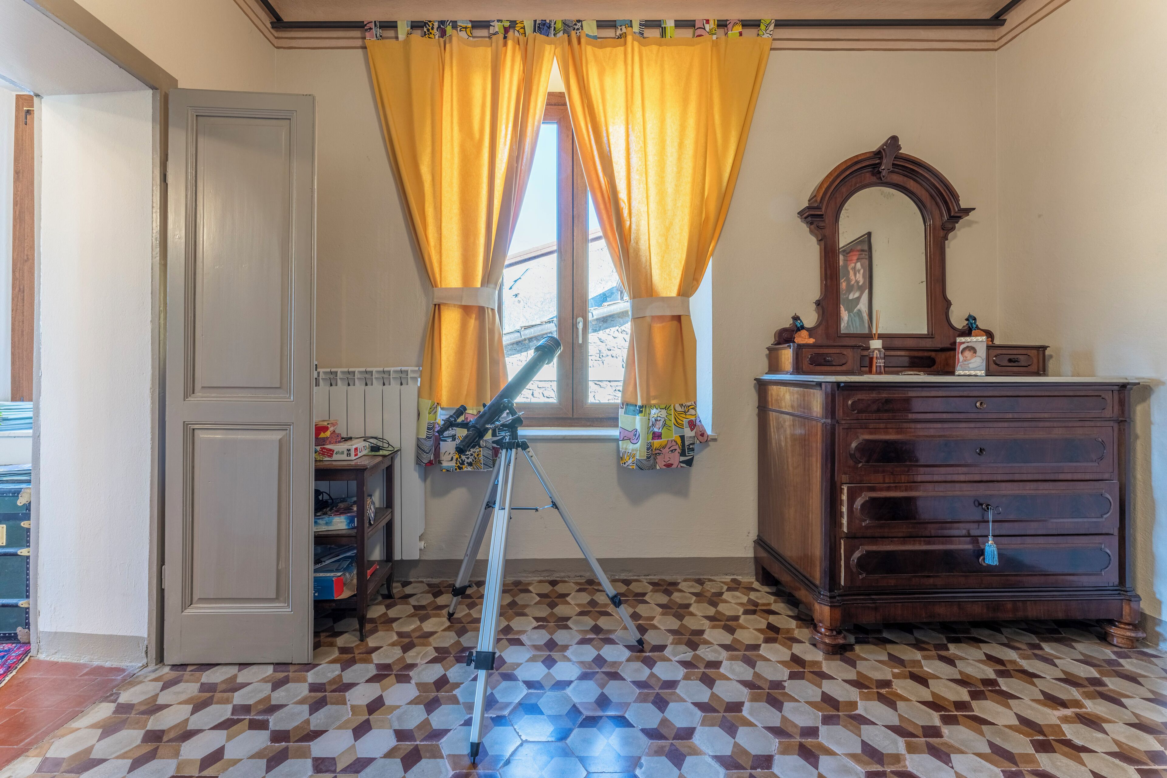 A room with a checkered floor, a wooden dresser, a tripod, and a window with yellow curtains.