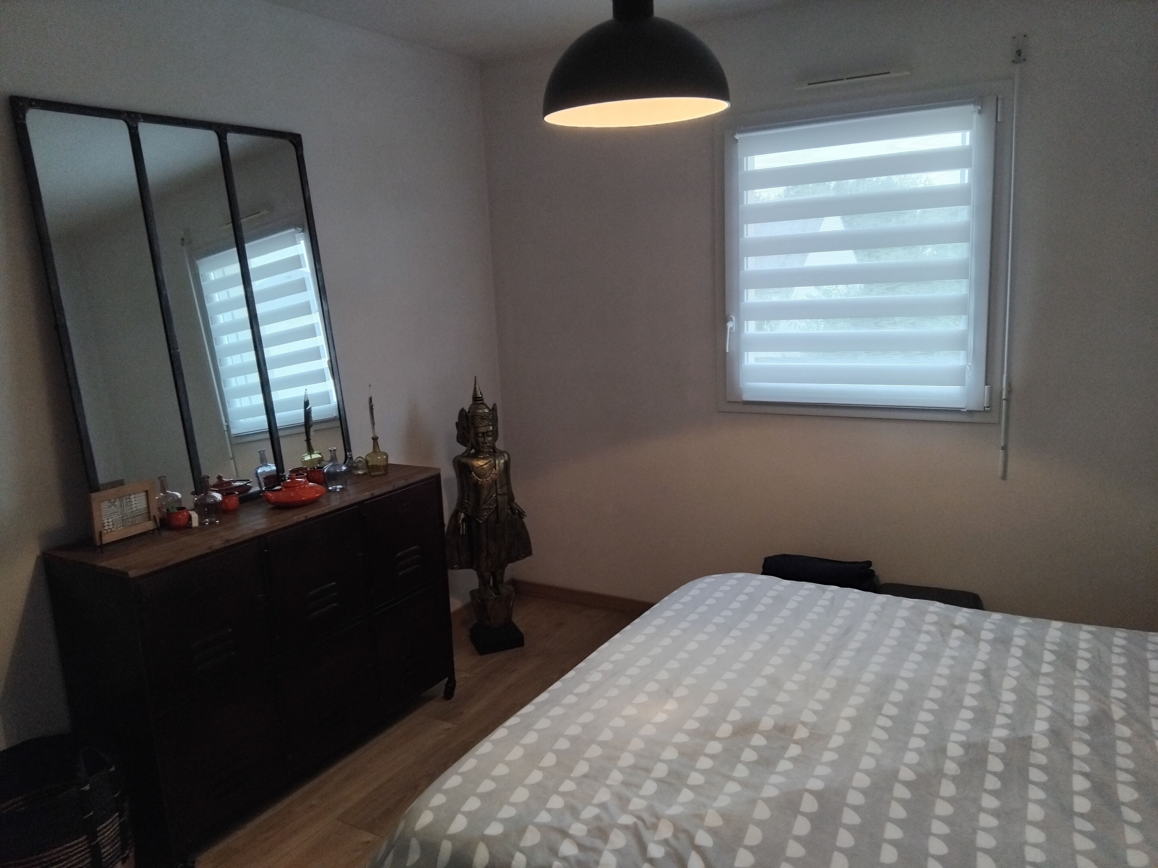 Een slaapkamer met een bed, een dressoir, een spiegel, een raam en een beeld.