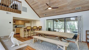 A spacious living area with a wooden ceiling, a dining table with benches, a kitchen in the background, and a large sliding glass door leading to a balcony with a view of trees.