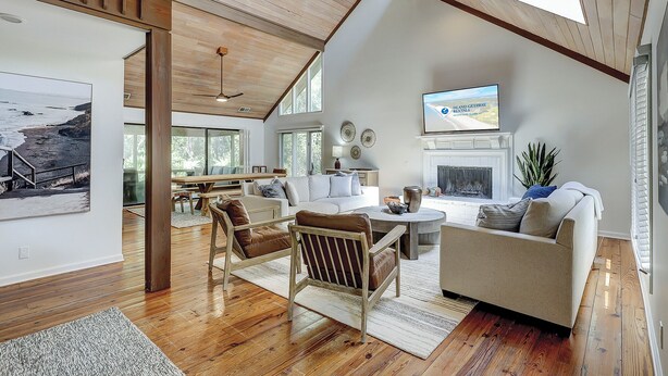 A spacious living room with a high ceiling, wooden beams, a fireplace, and a large TV screen.