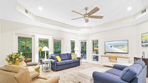 A spacious living room with a ceiling fan, large windows, a flat-screen TV, and a comfortable seating area with a sofa and chairs. - Luxury Wendover Dunes villa, steps to ocean (Hilton Head Island)