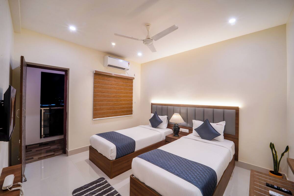 A hotel room with two beds, a ceiling fan, an air conditioning unit, a bedside table with a lamp, and a small potted plant.
