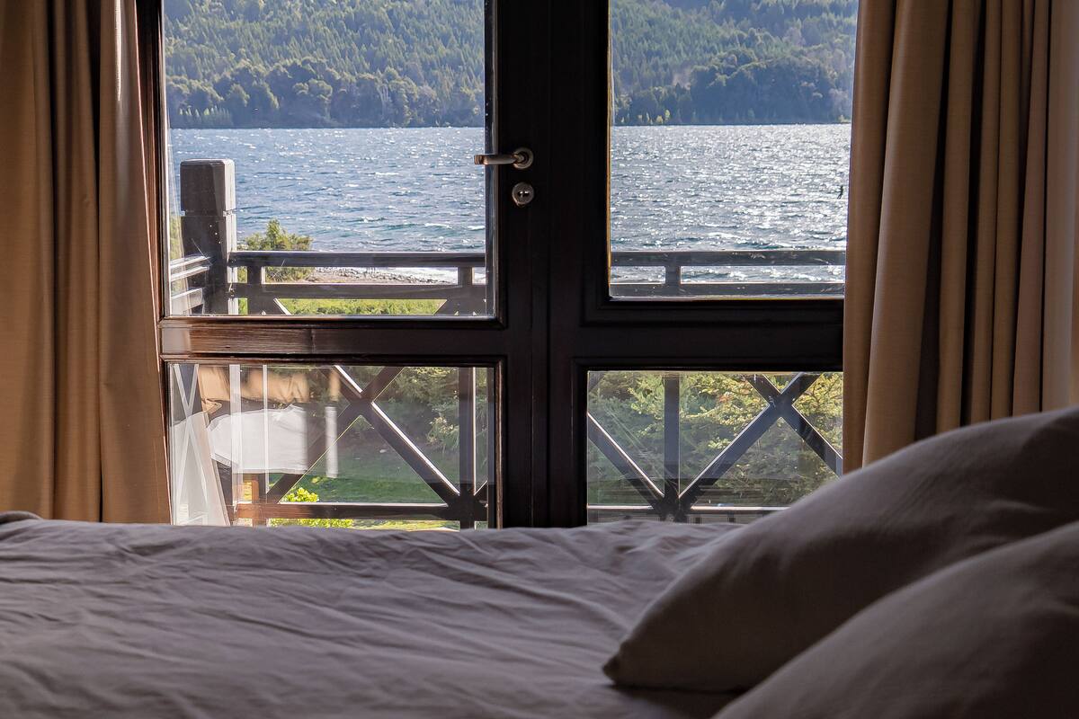 Una cama con ropa de cama blanca, vista a un lago con una colina boscosa al fondo y un balcón con barandilla.