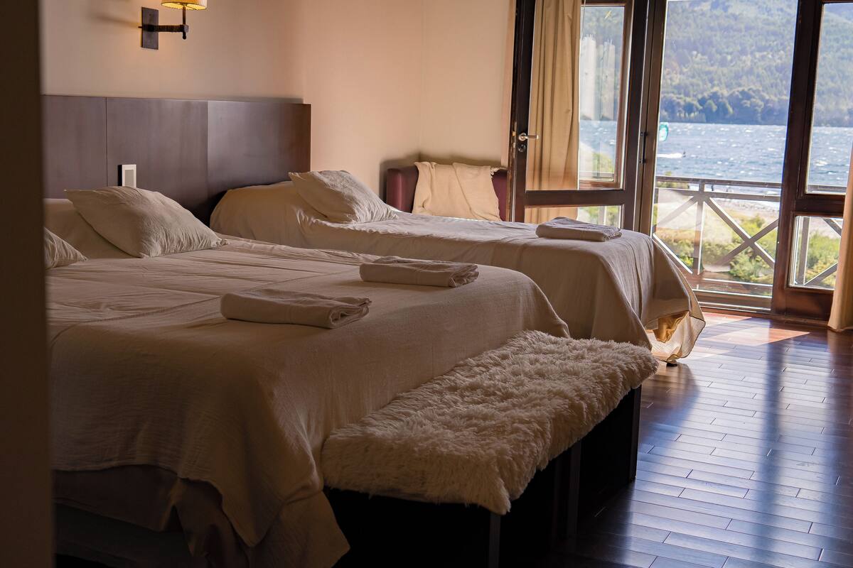 Habitación de hotel con dos camas, un banco y vista al mar a través de un amplio ventanal.