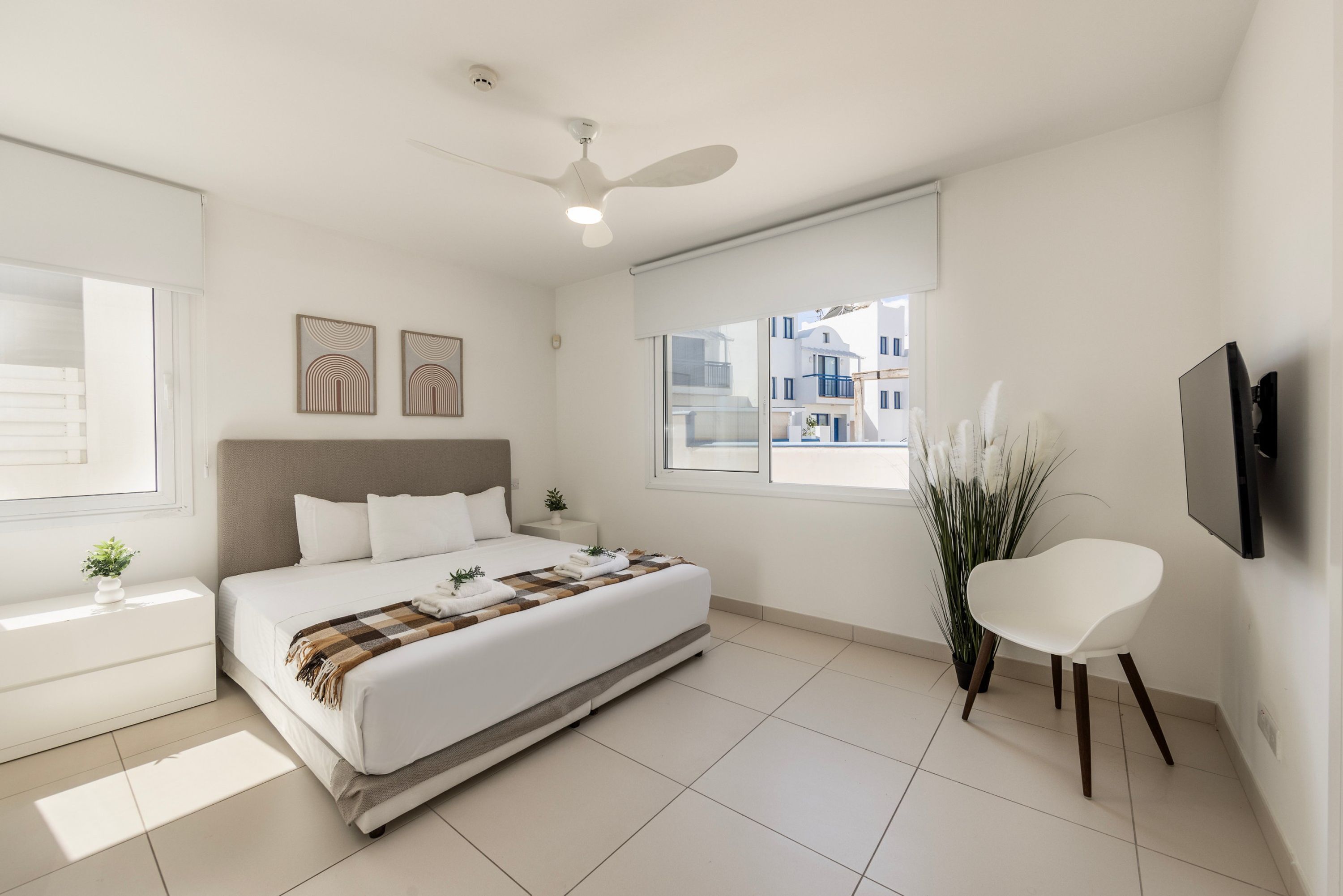 Un dormitorio moderno con cama, mesitas de noche, ventilador de techo y ventana con vistas a edificios.