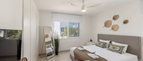 A modern bedroom with a large bed, a ceiling fan, a window with a view, and a small bedside table.