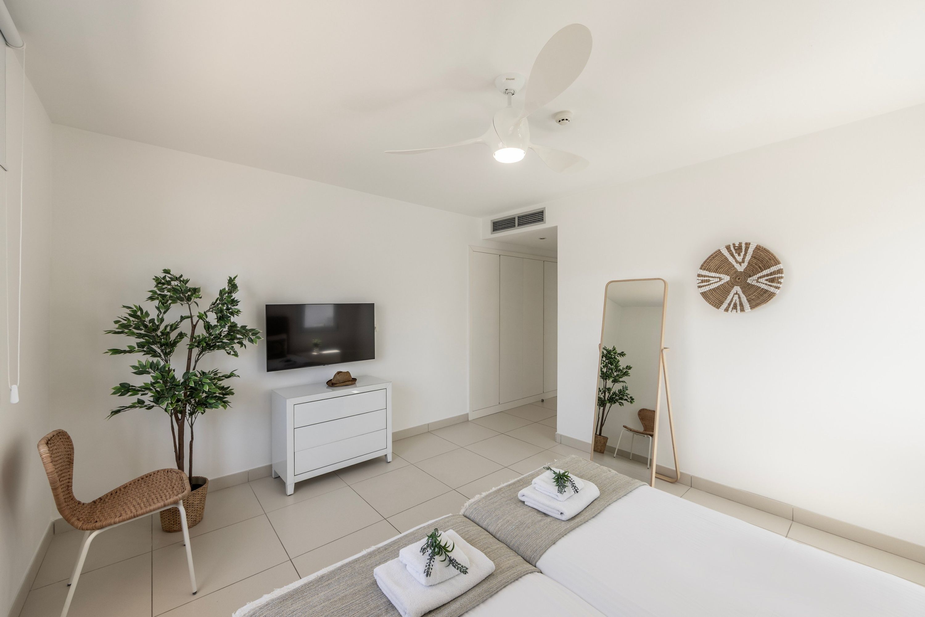A modern bedroom with a bed, a TV, a ceiling fan, a white dresser, a chair, and two potted plants.