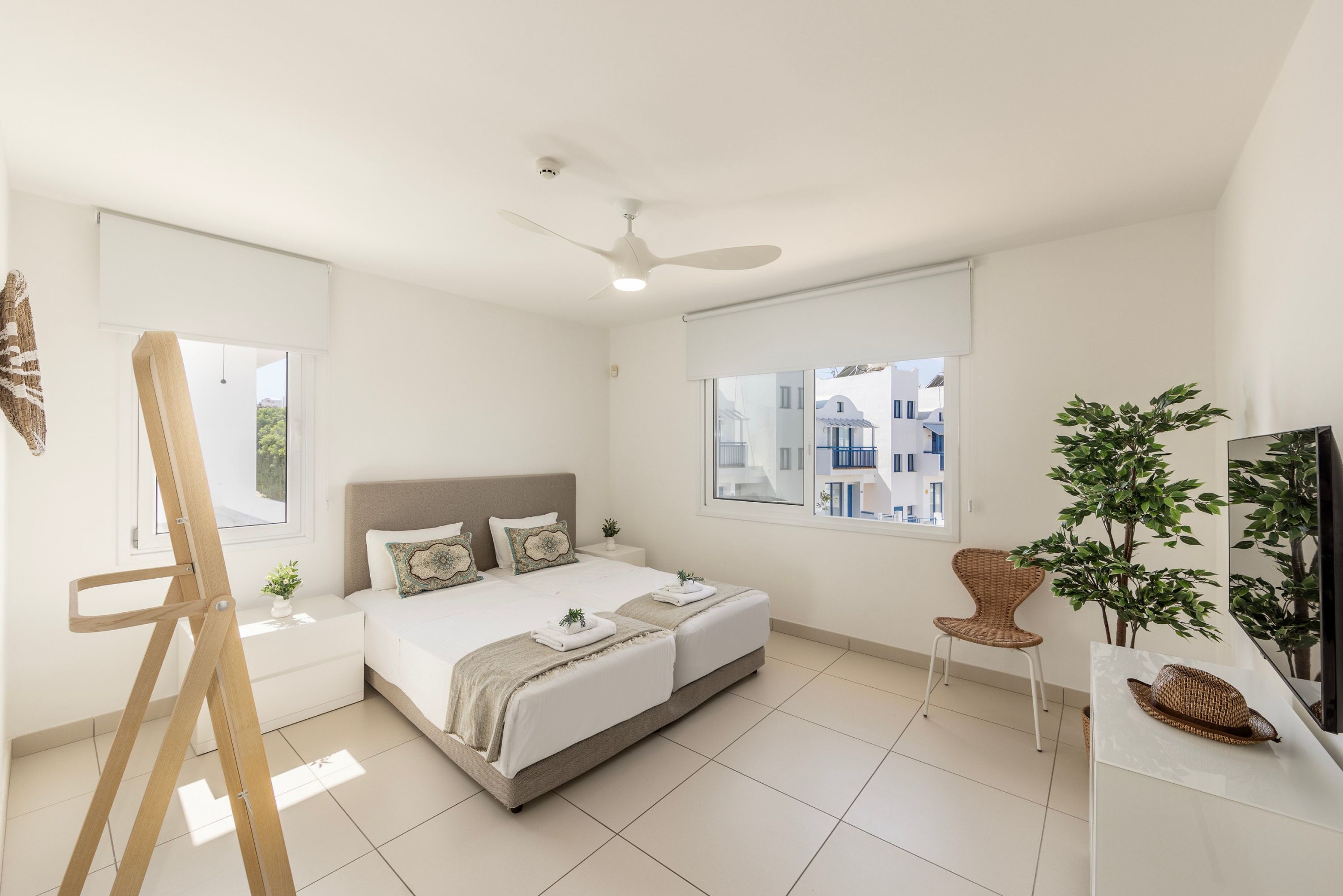 A modern bedroom with a large bed, a ceiling fan, a TV, a chair, and a window with a view of buildings.