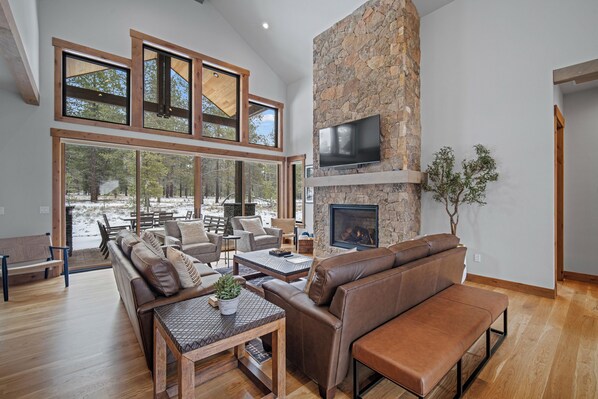 A spacious living room with a stone fireplace, leather sofas, and large windows overlooking a snowy landscape. - Luxury Family Escape with Access to Exclusive Golf (Sunriver)
