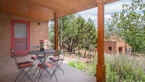 Une terrasse extérieure avec une table et des chaises, une porte rouge et, en arrière-plan, une vue sur des arbres et un bâtiment.