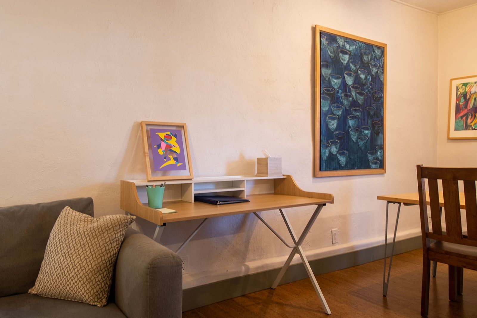 A living room with a sofa, a wooden desk, and two chairs. There are framed artworks on the wall.